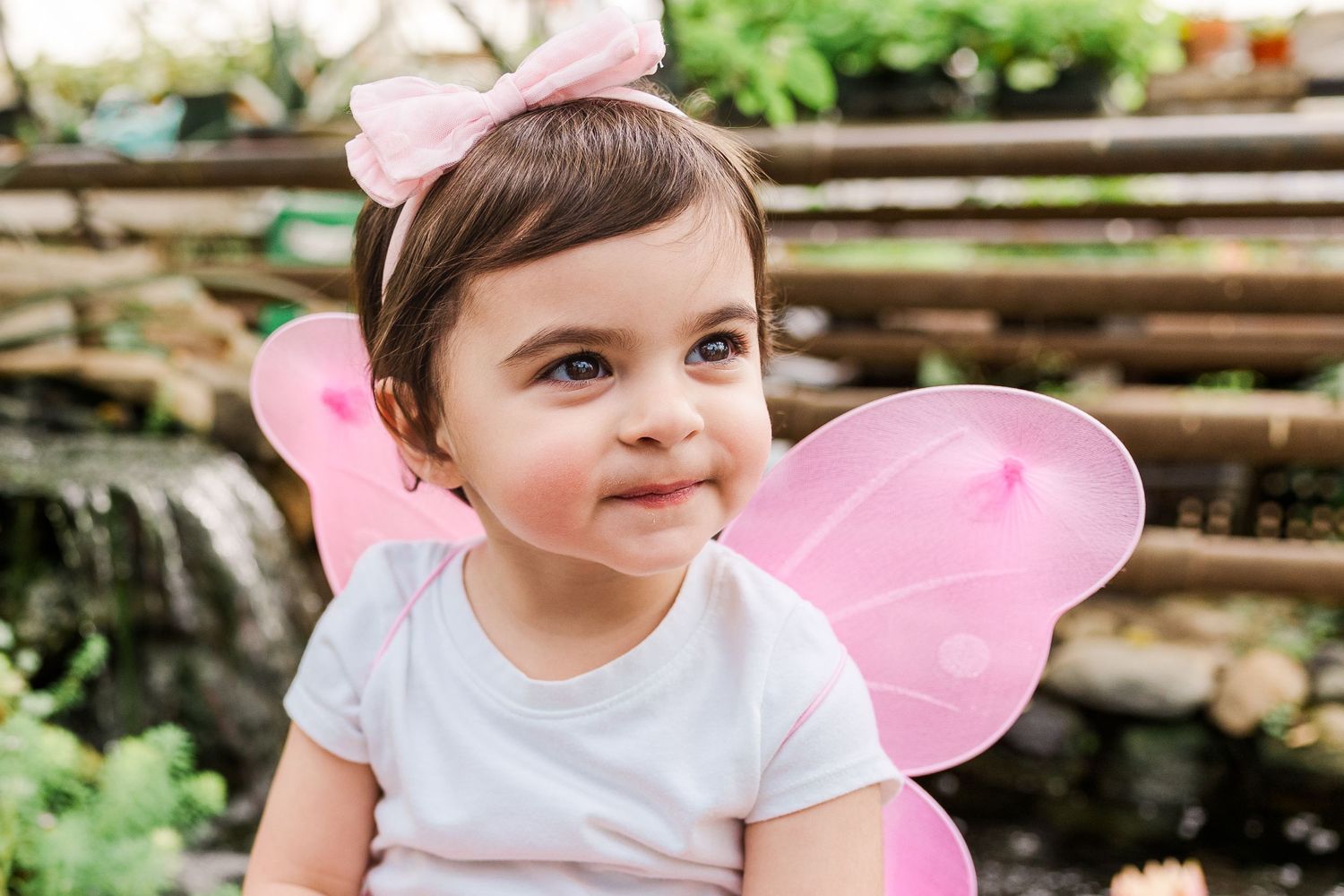 Happy toddler wearing pink fairy wings and bow headband smiles in garden setting.