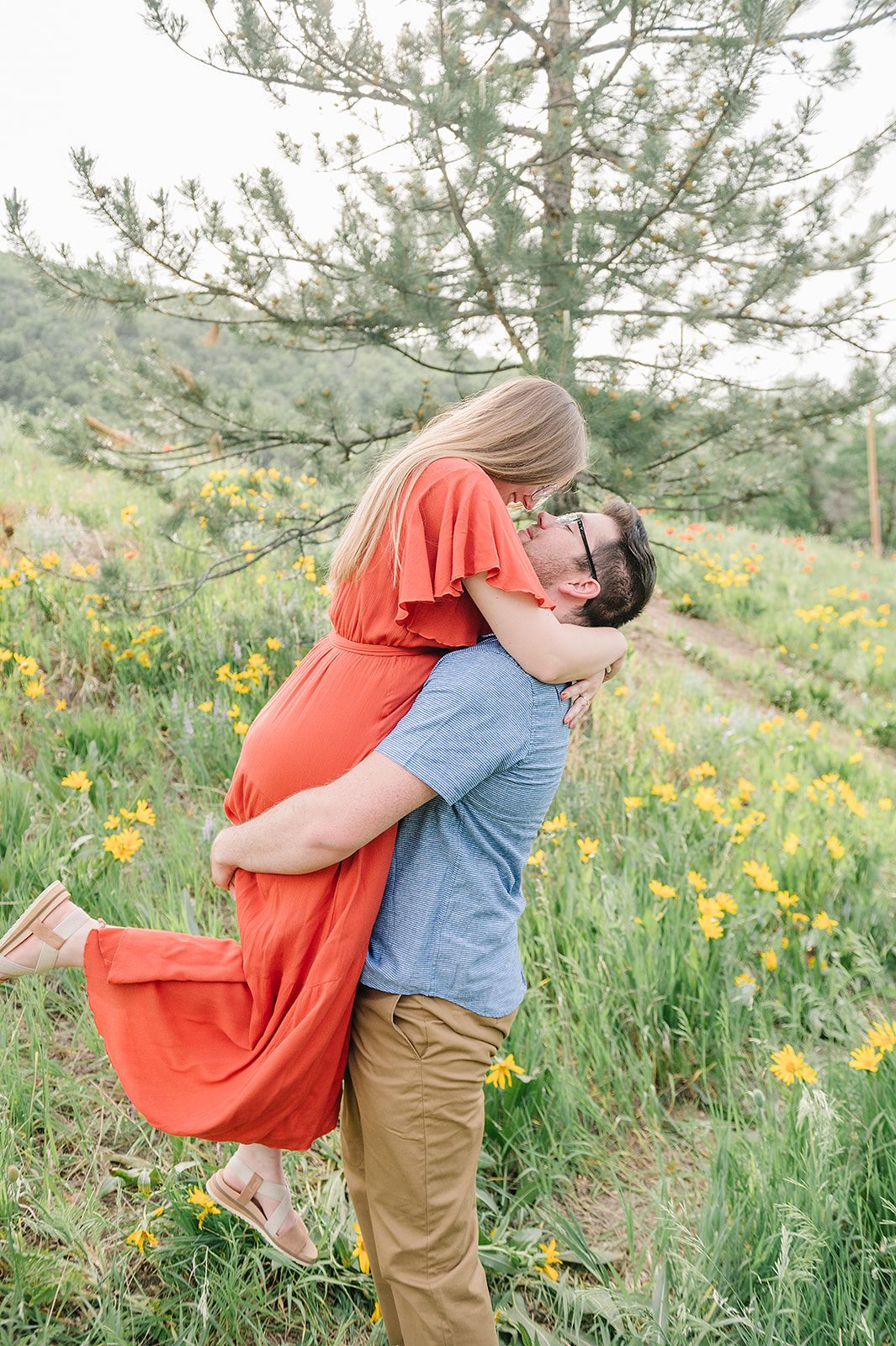 Romantic lift pose in a meadow filled with yellow wildflowers and pine trees.