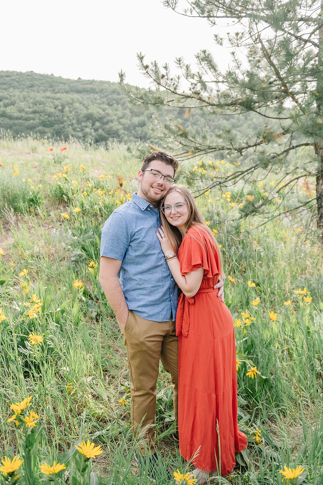 A couple embraces in a meadow filled with yellow wildflowers and mountain scenery.
