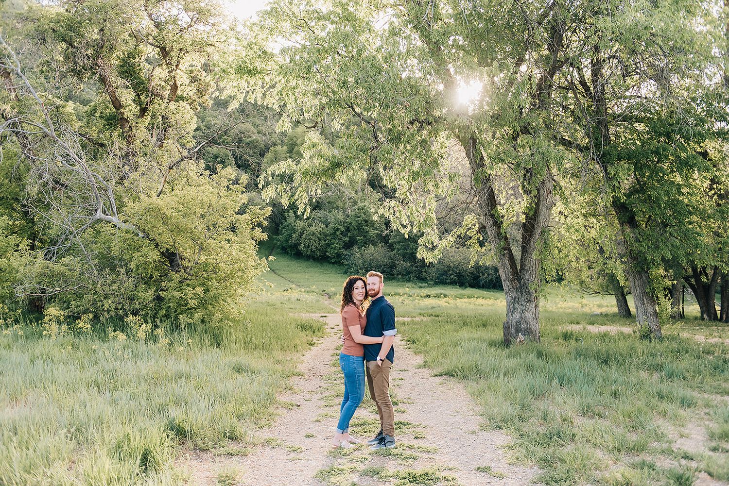 Couple embracing on a sunlit dirt path surrounded by oak trees and green grass.