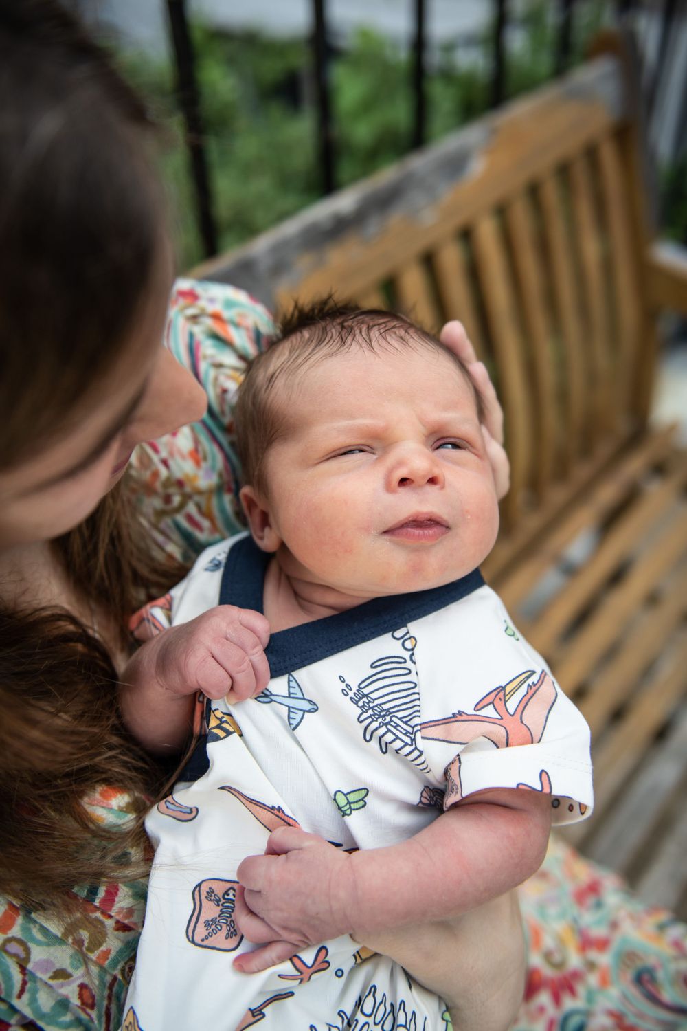 Newborn baby in animal print onesie looking up while being held outdoors.