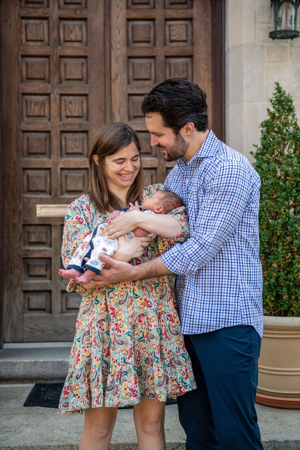 Family stands by wooden door as parents cradle newborn baby wearing white clothing.