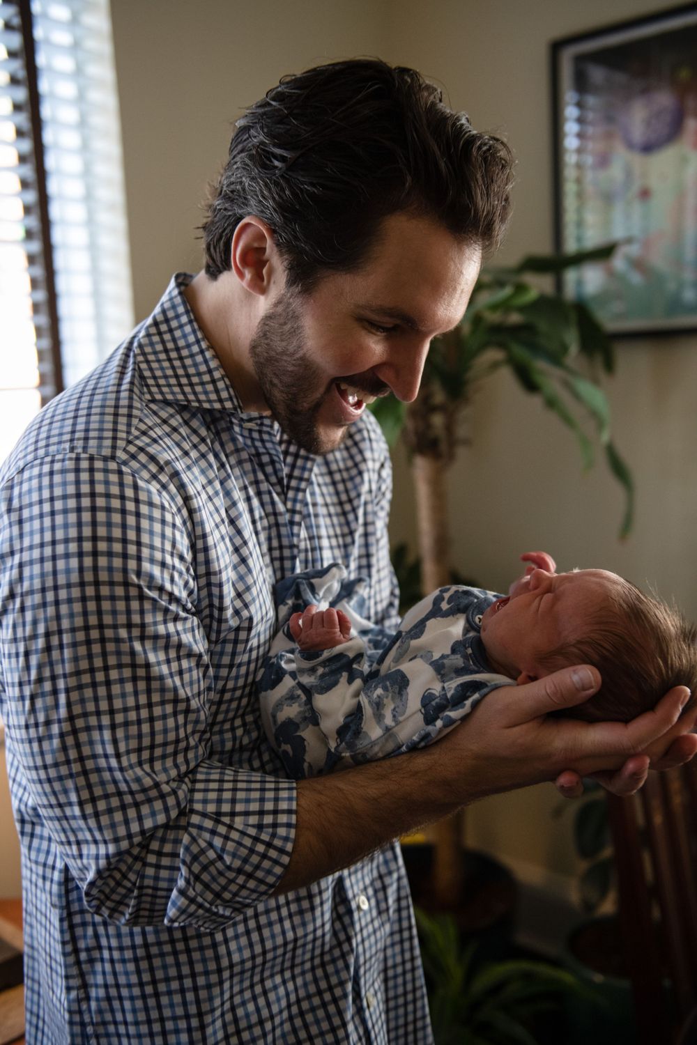 Baby wearing blue patterned sleeper smiles while being held in indoor setting.