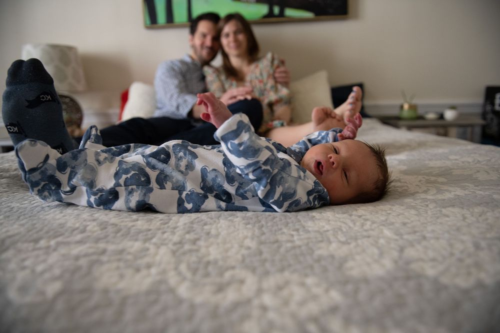 Baby in blue patterned sleeper lies on bed with soft focus background.
