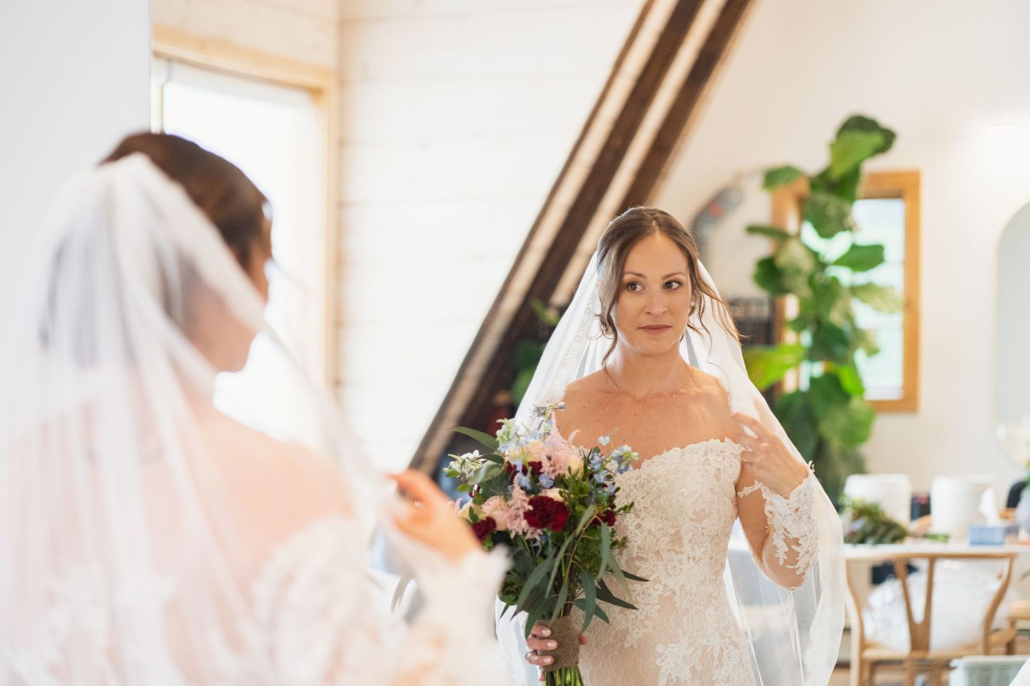 Bride in an elegant lace wedding dress admires her reflection while holding a burgundy and white bouquet.