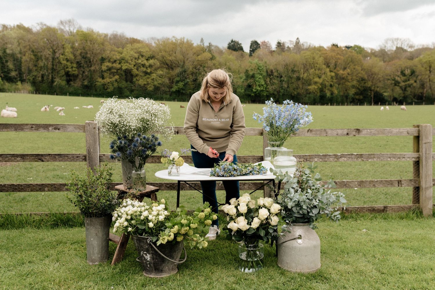 Florist arranging fresh flowers at an outdoor table against a pastoral countryside backdrop.