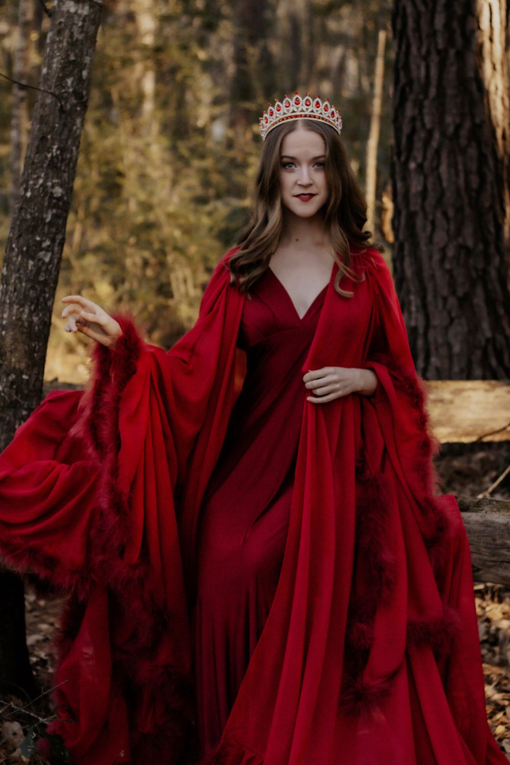 A woman in a red dress and crown stares into the camera while standing in the woods