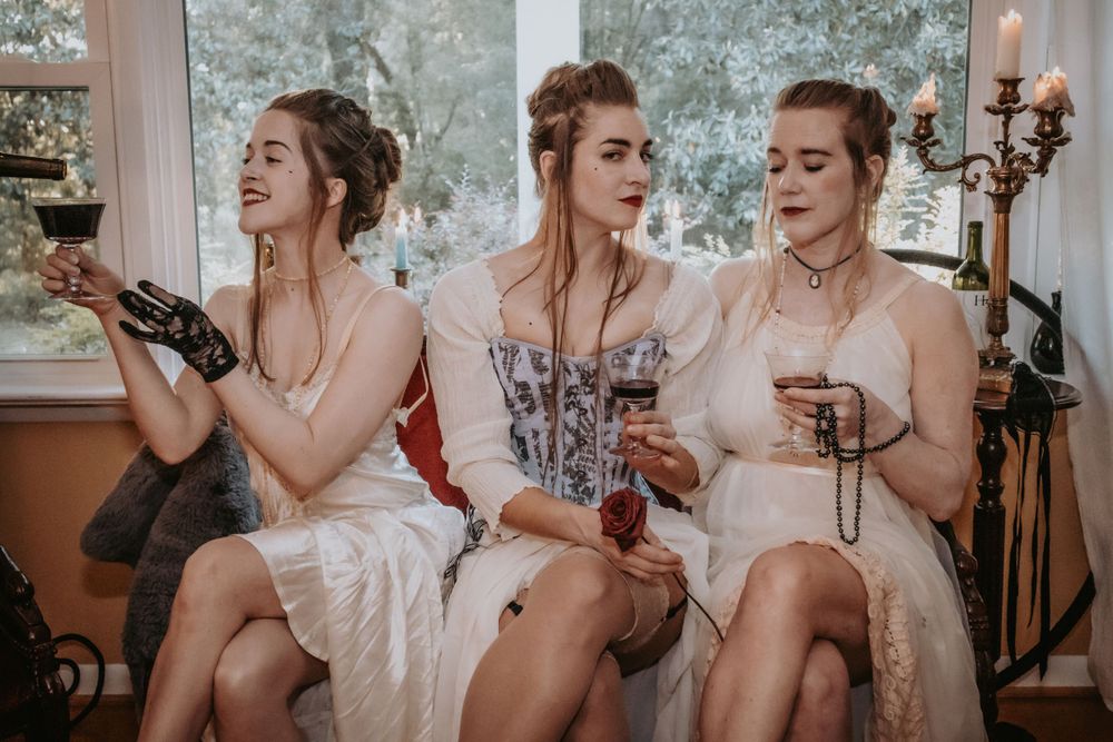Three women in 1800s style clothing pass a glass of wine between each other