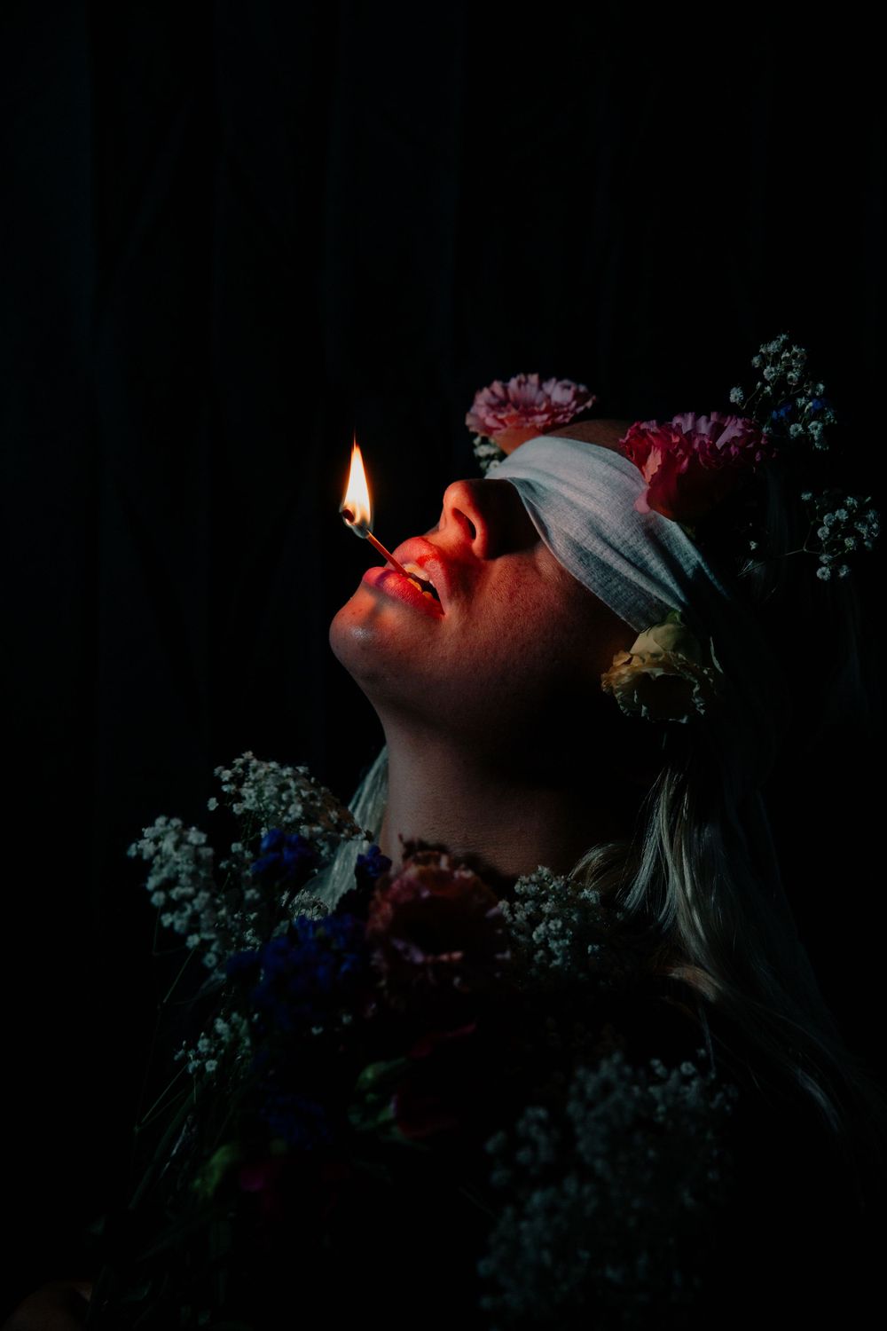 A woman poses in darkness surrounded by flowers while holding a lit match in her mouth
