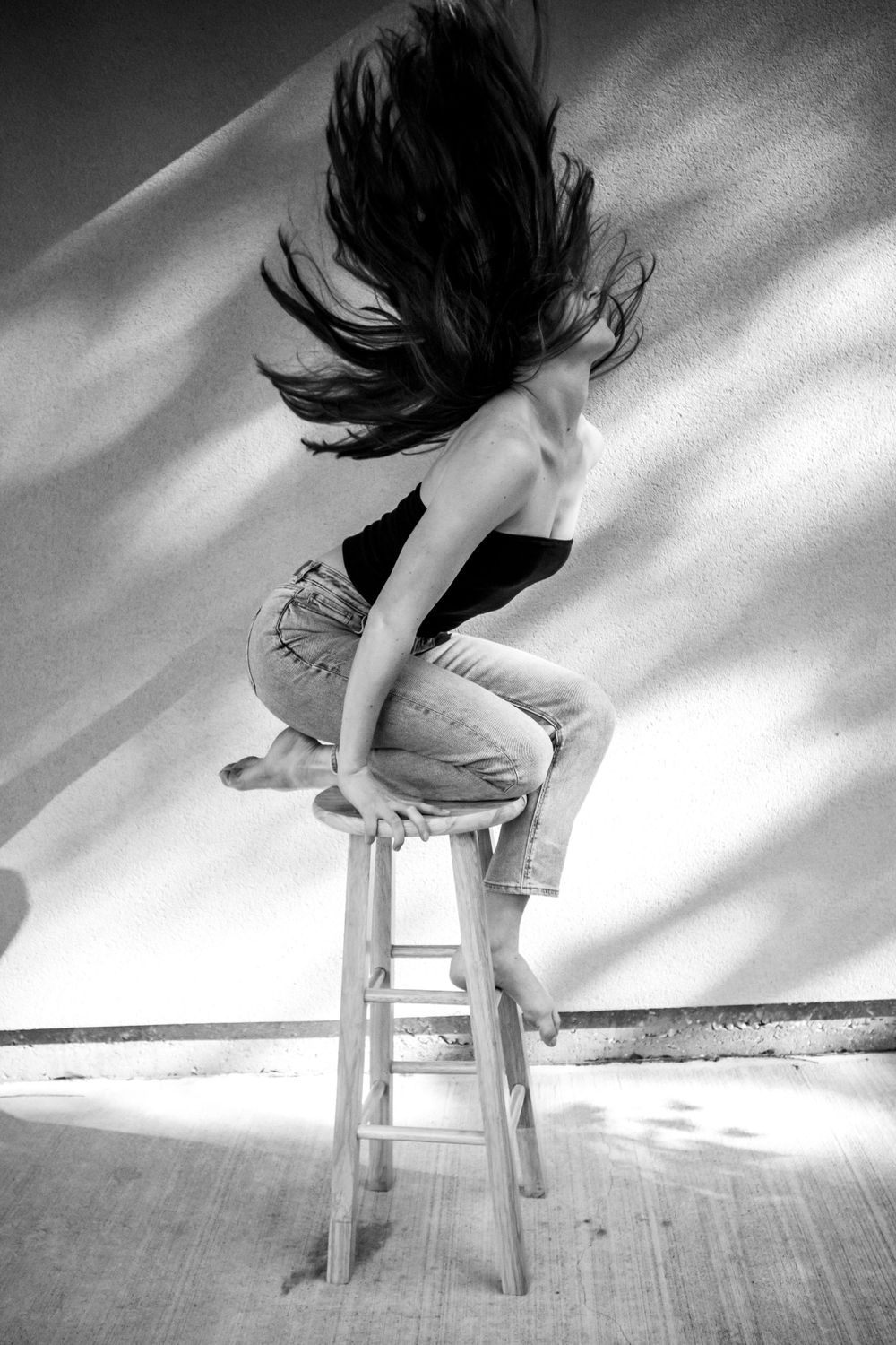 A black and white photo of a woman tossing her hair back while seated on a stool