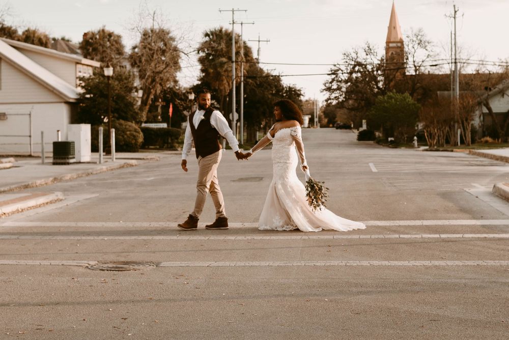 Newlyweds walk hand in hand down a sunset-lit street with a church steeple in the background.
