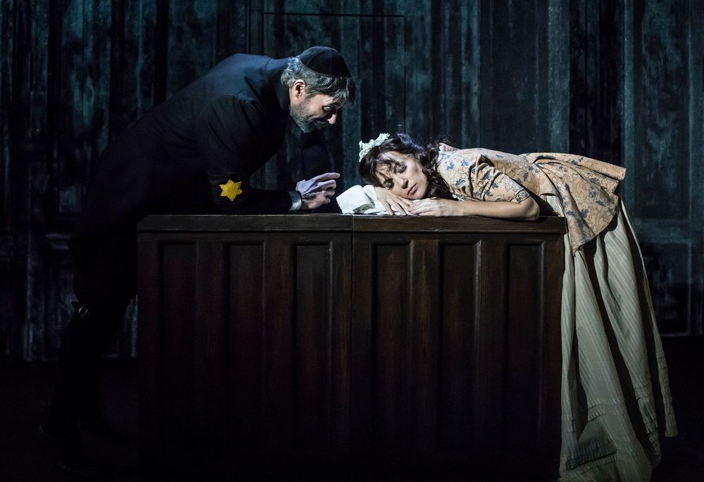 A dramatic scene on a dark theater stage with performers at a wooden table.