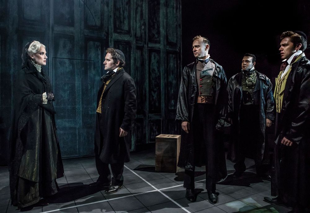 Gothic stage scene with performers in dark period costumes gathered in dramatic confrontation.
