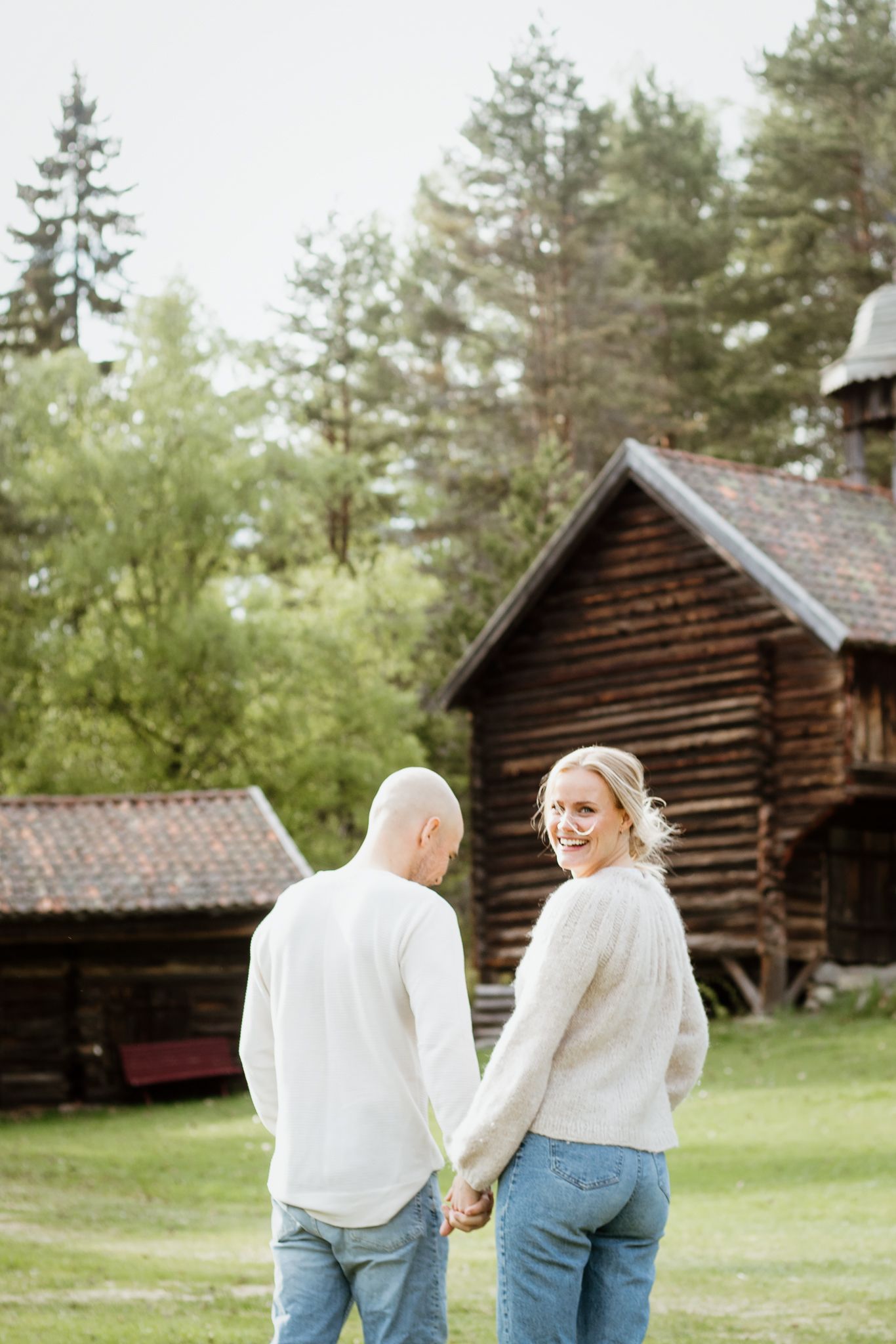 Couple holding hands while walking near rustic log cabins in forest setting.