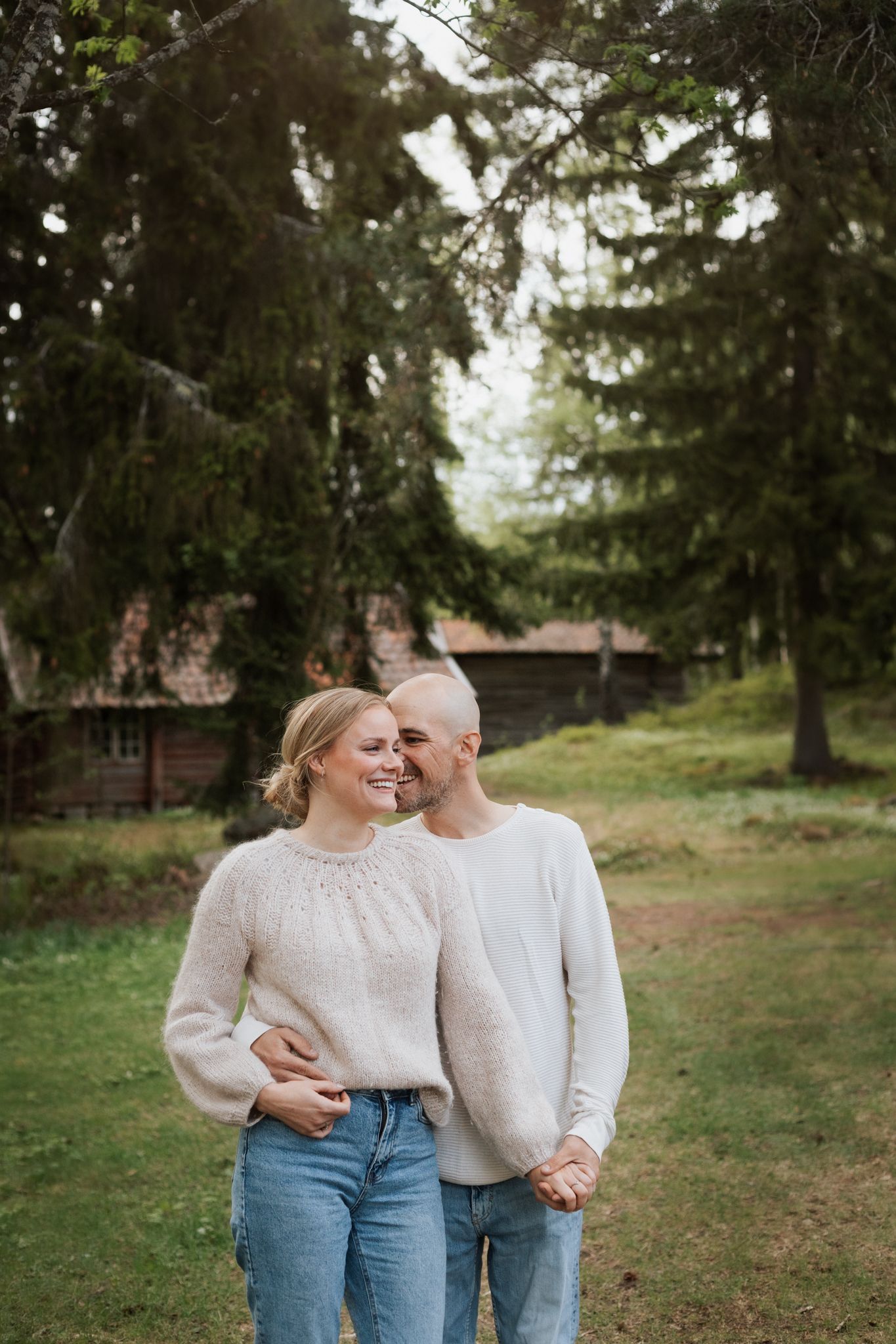 Couple in matching cream sweaters walking together near rustic cabin in pine forest.