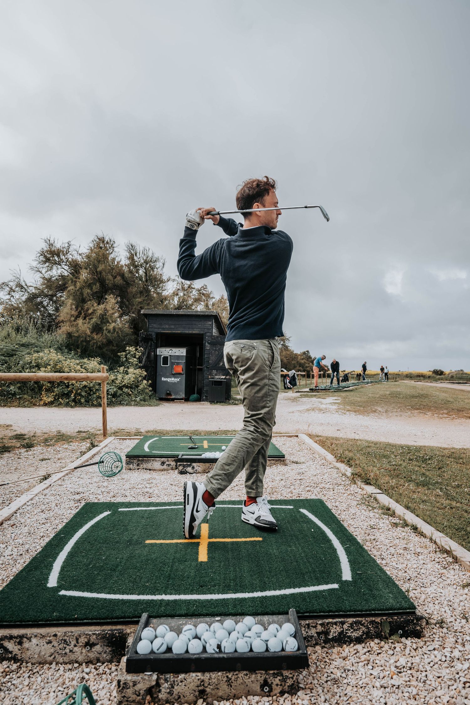Golfer practicing swing on artificial turf mat with golf balls lined up at outdoor driving range.