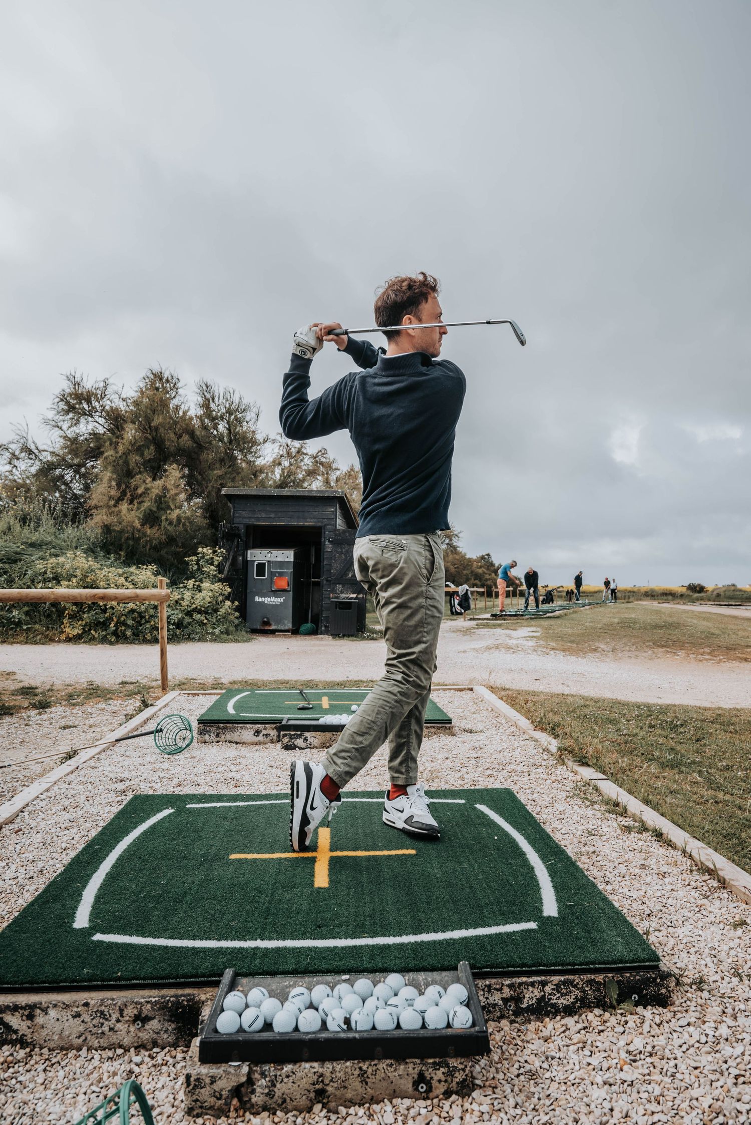 Golfer following through swing stance on practice mat at outdoor driving range under cloudy sky.