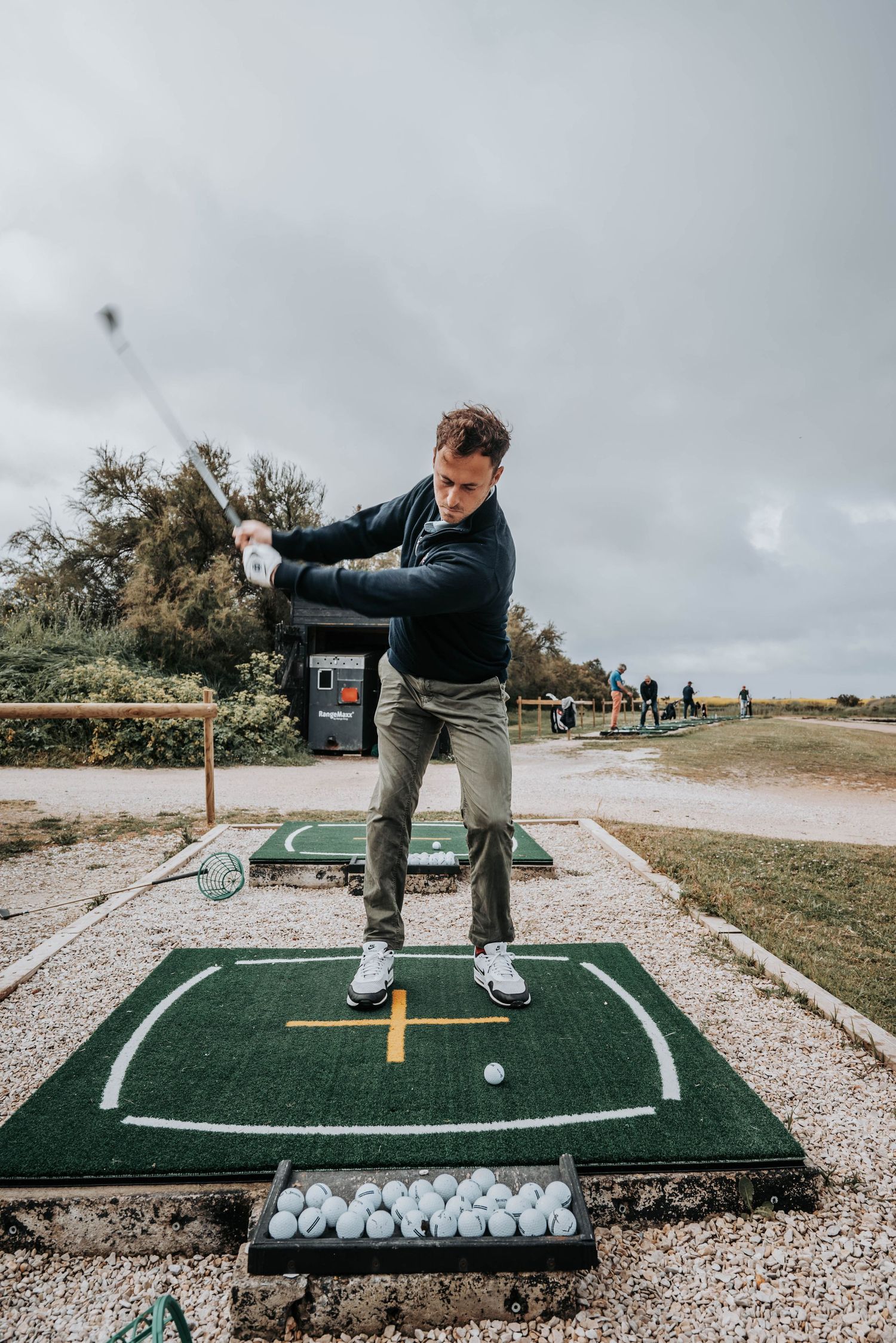 Golfer mid-backswing at an outdoor practice facility with cloudy sky backdrop.