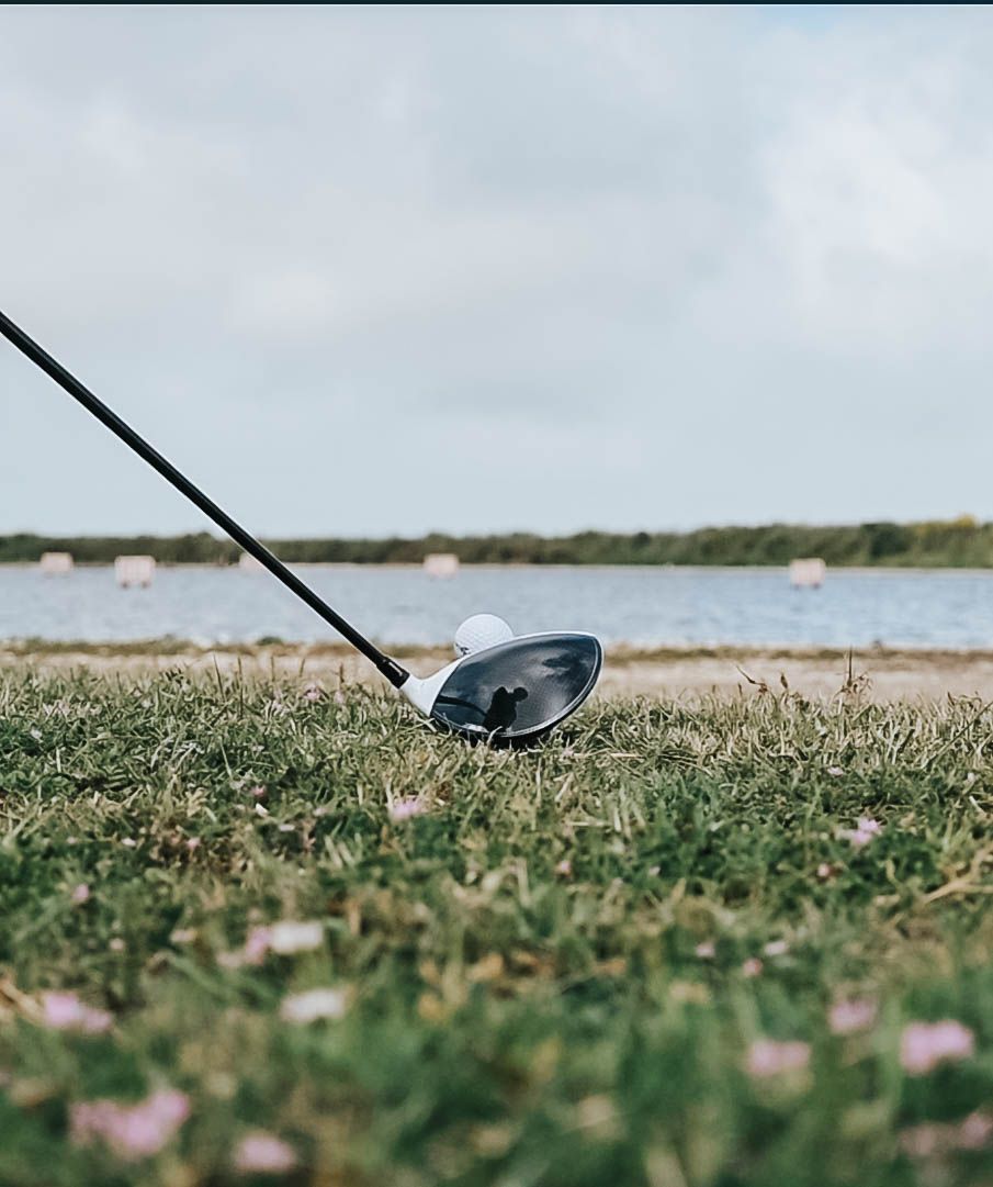 Close up of golf driver club head resting on grass with water hazard in background.