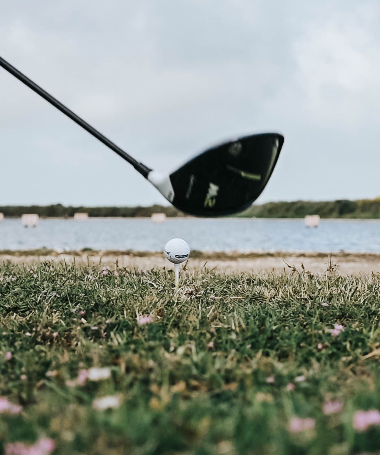 Golf club head positioned behind white golf ball on grassy course with water in distance.