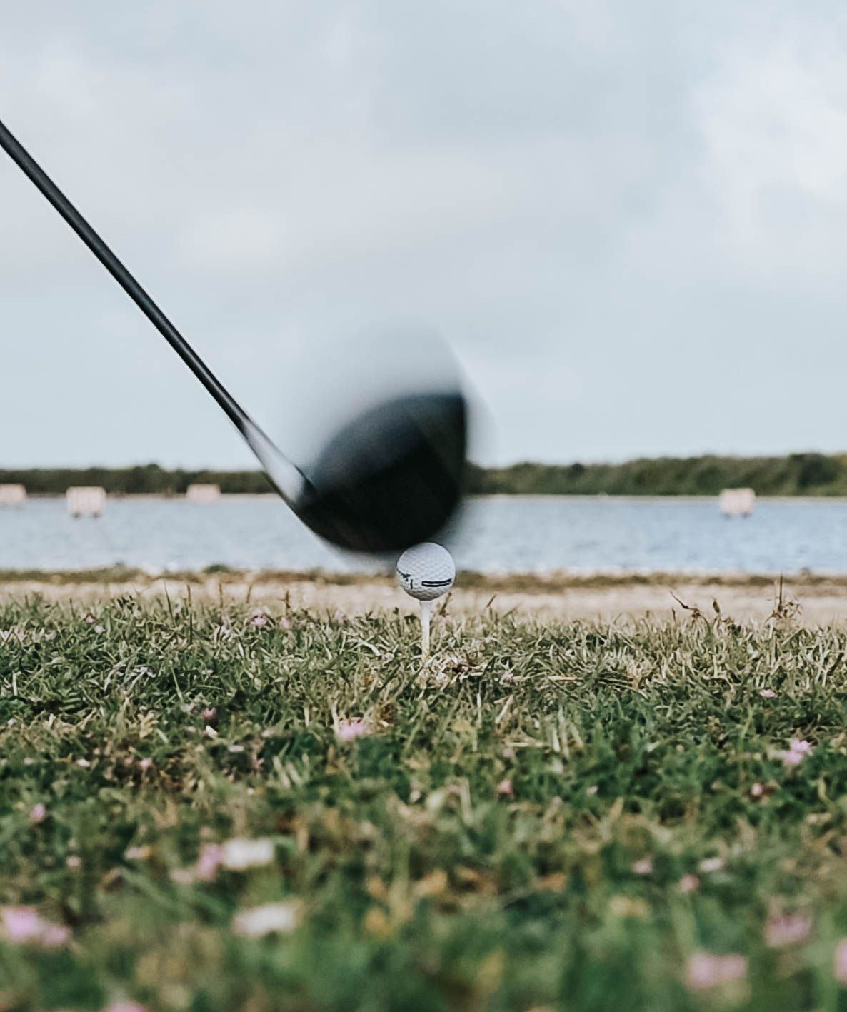 Golf club positioned to strike ball on tee with lake and shoreline visible in background.