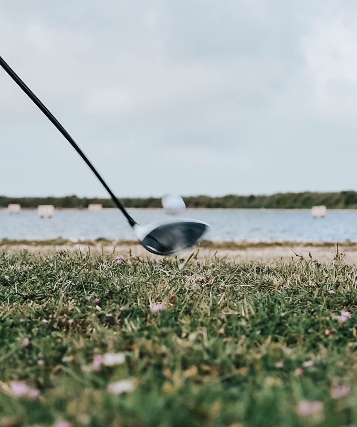 Golf club making contact with ball on tee against waterfront course backdrop.