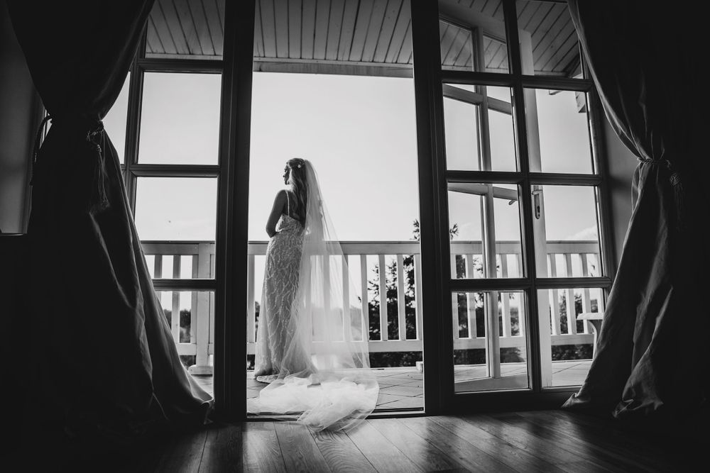 Silhouette of a bride in flowing white dress standing on balcony in dramatic backlit window scene.