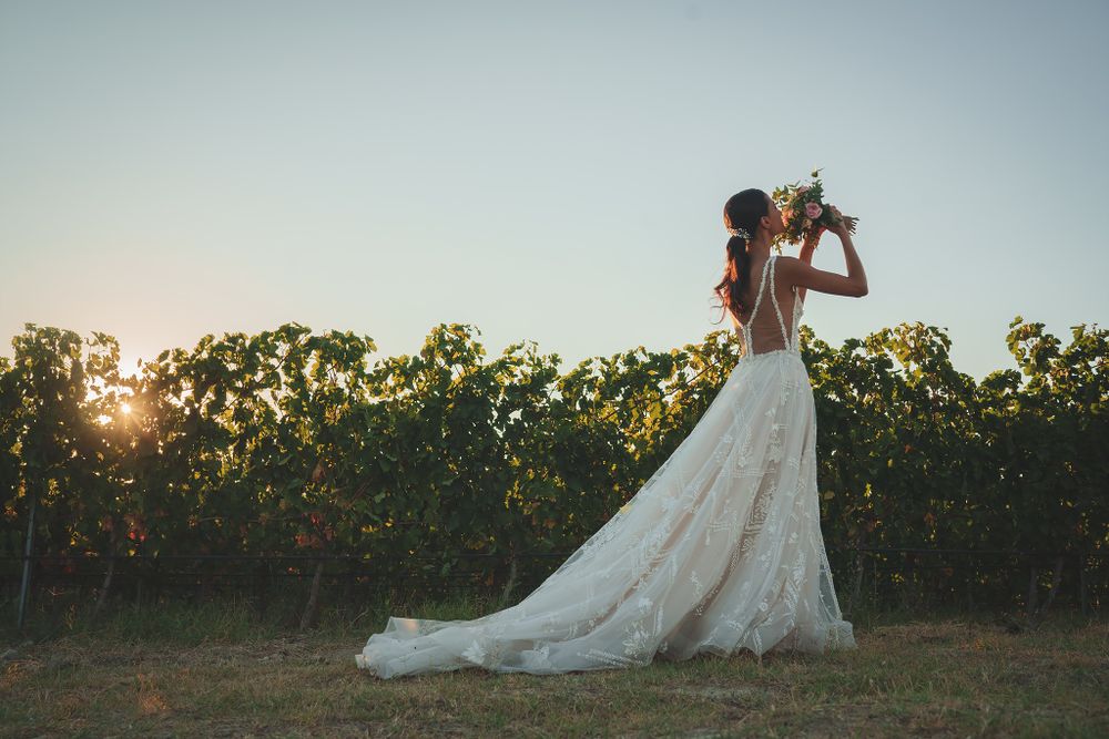 A bride in a flowing lace dress poses in a vineyard at sunset.
