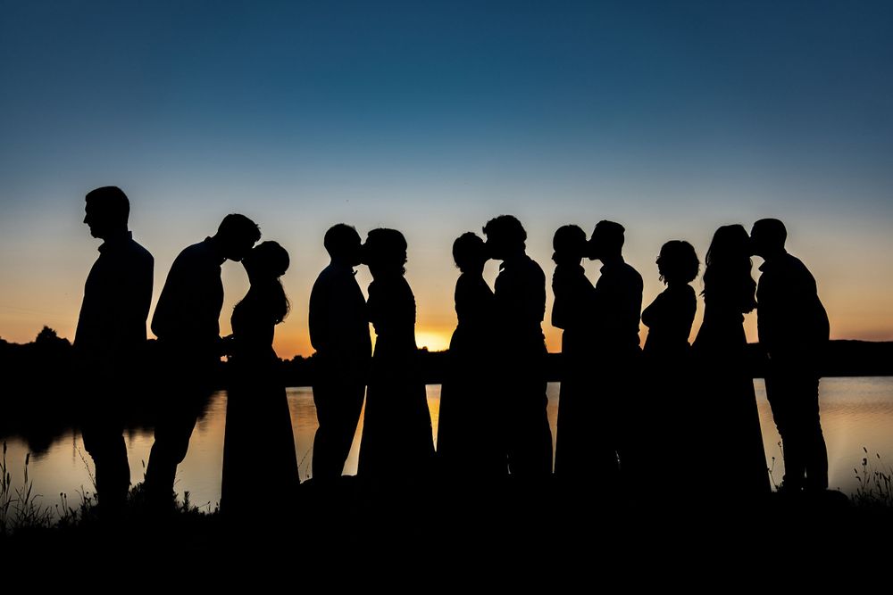 Silhouetted wedding party poses against sunset sky by a lake.