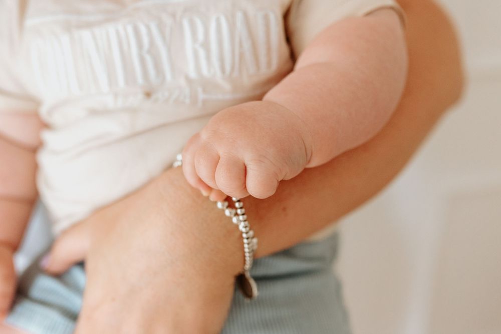 Close up of baby's hand wearing delicate beaded bracelet against light colored clothing.