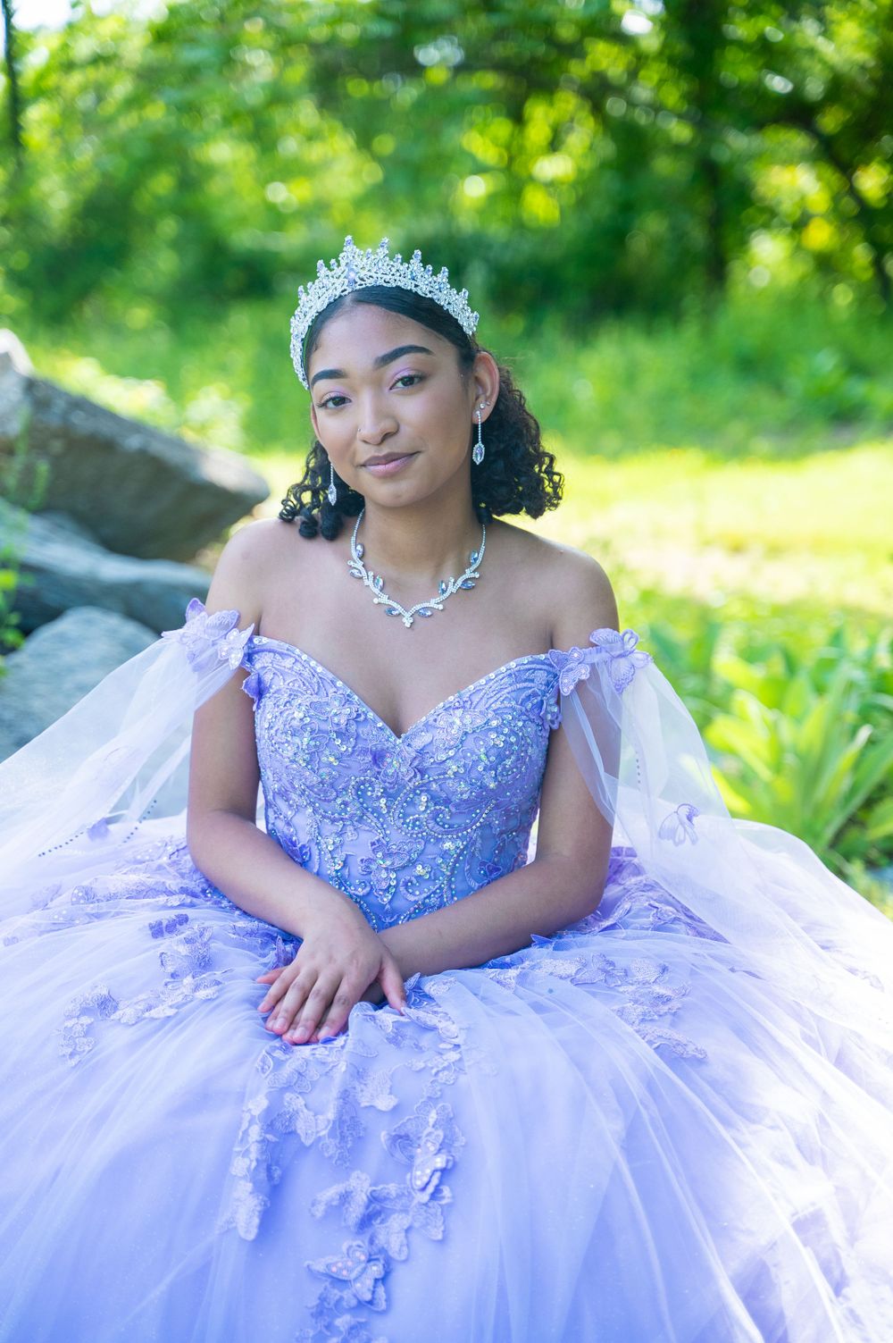 A stunning lavender ball gown with off-shoulder design, crystal tiara and matching jewelry set against a nature backdrop.