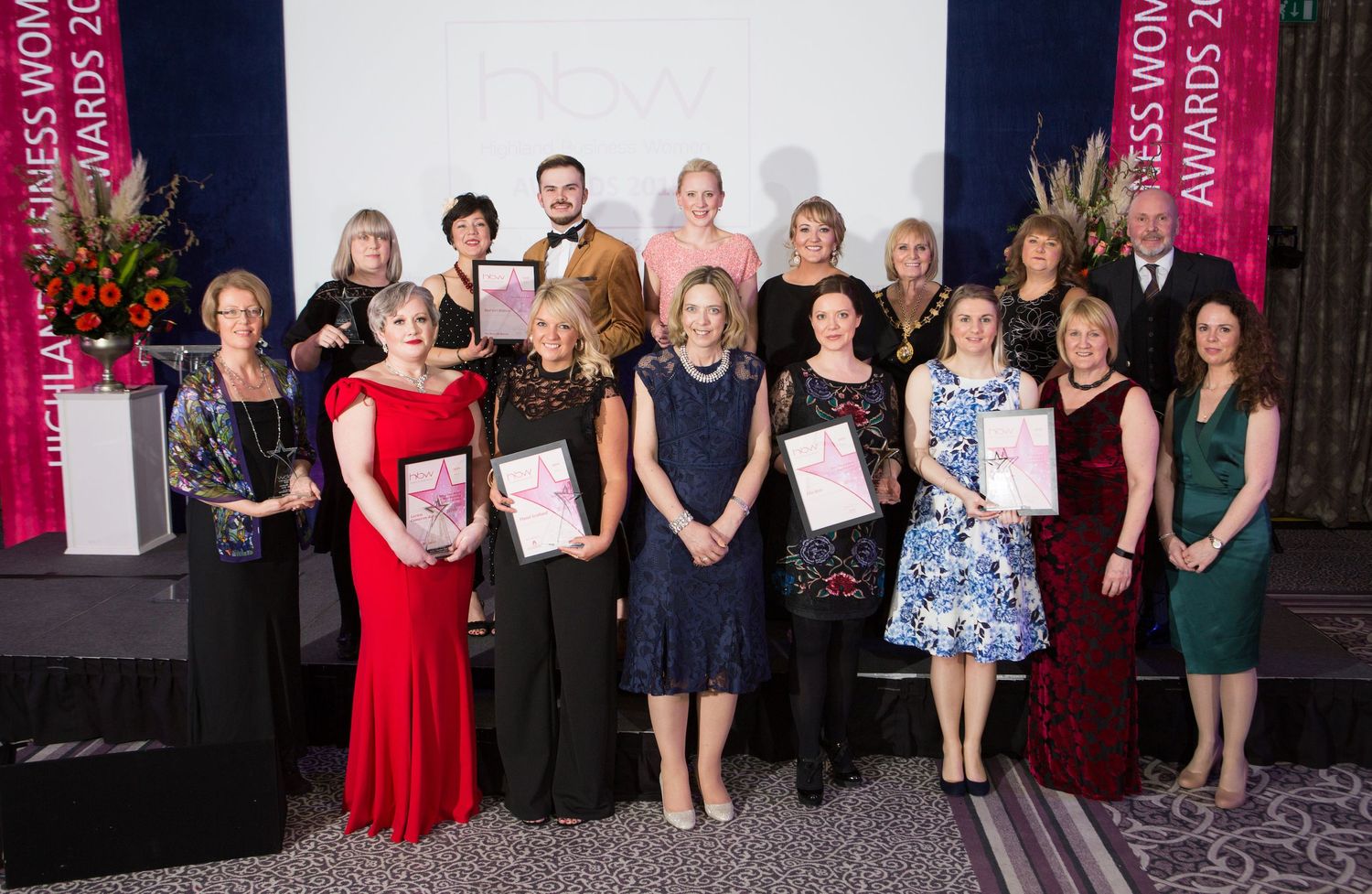 Group photo of award recipients at a formal ceremony with red banners and floral arrangements.