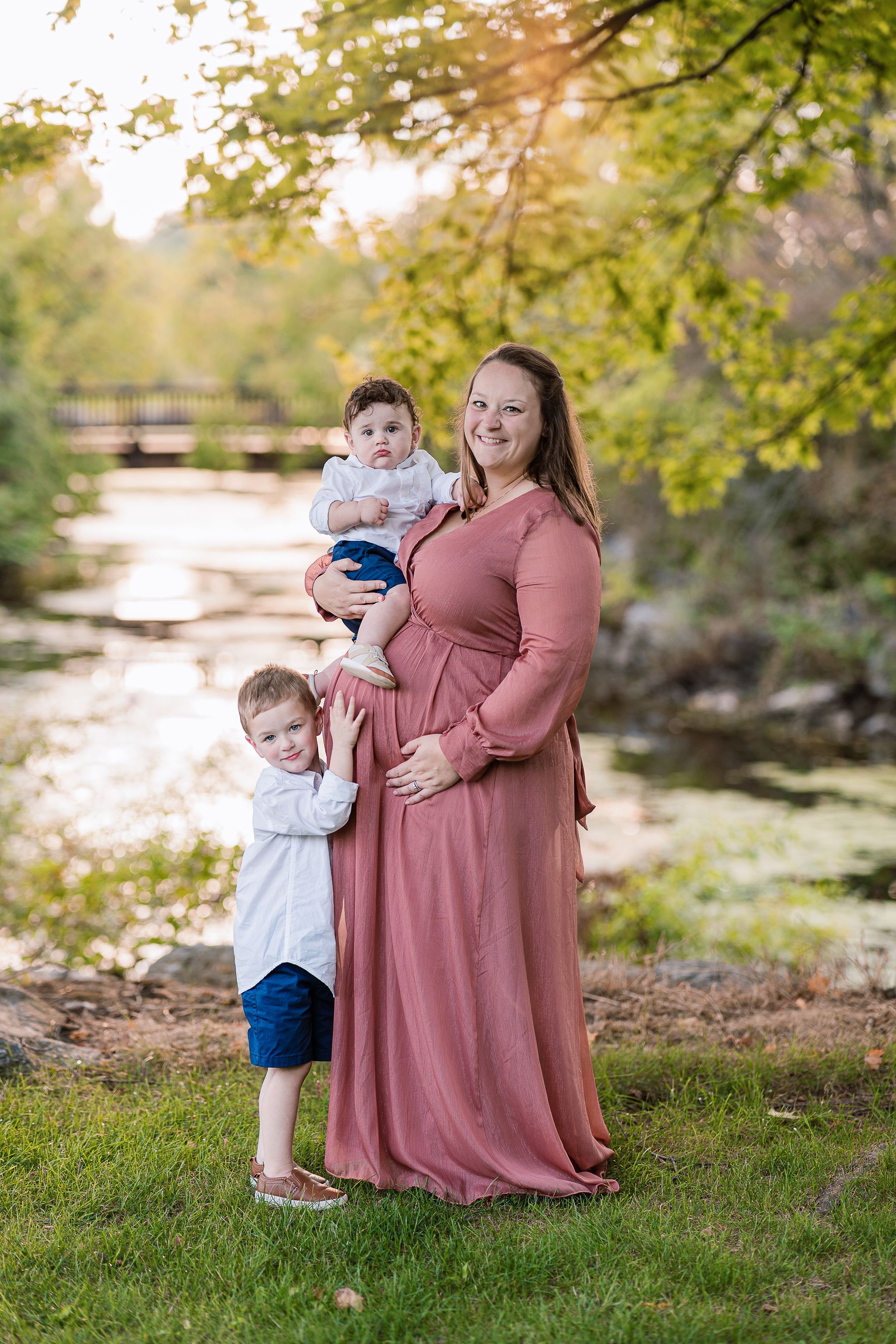 Expecting mother in pink dress poses with children by scenic pond.