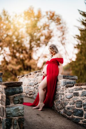 A pregnant woman in a red dress poses elegantly against a stone wall during golden hour.