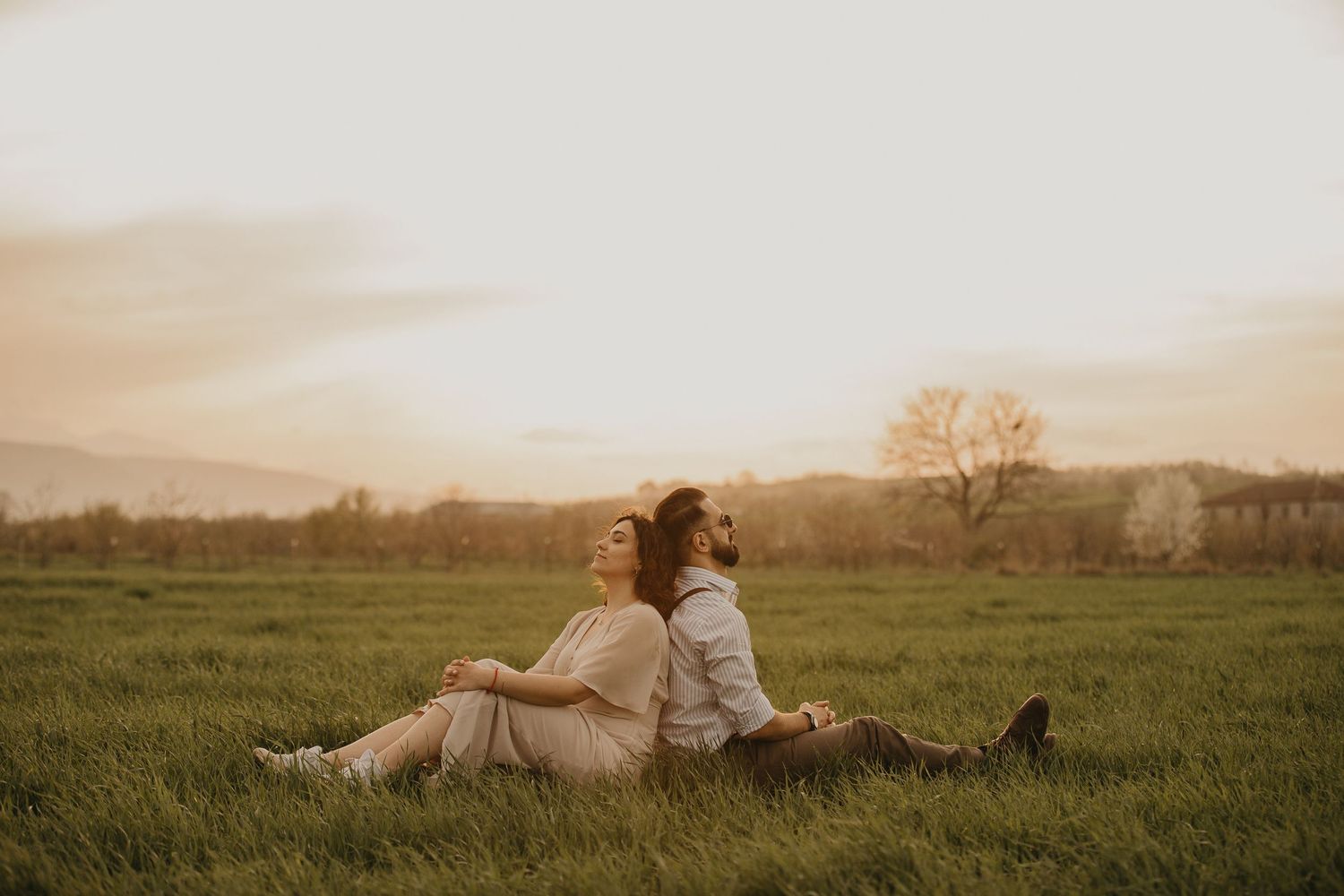 A couple rests together in a serene meadow at sunset with mountains in the distance.