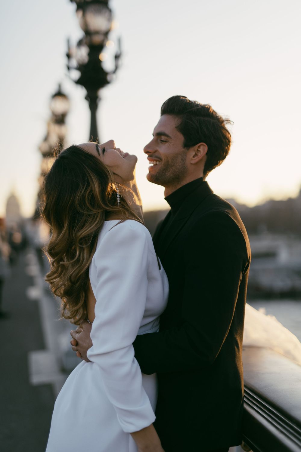Couple in black and white outfits embracing against sunset urban backdrop.
