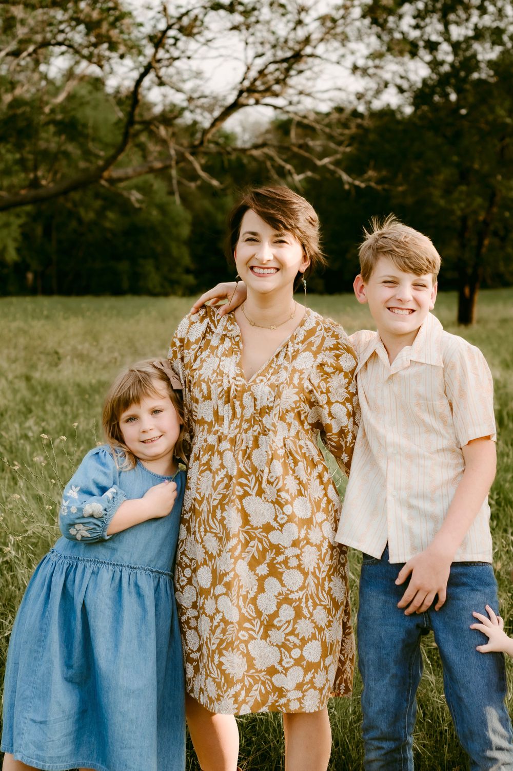 Three people pose for a casual outdoor portrait in a meadow with trees in the background.