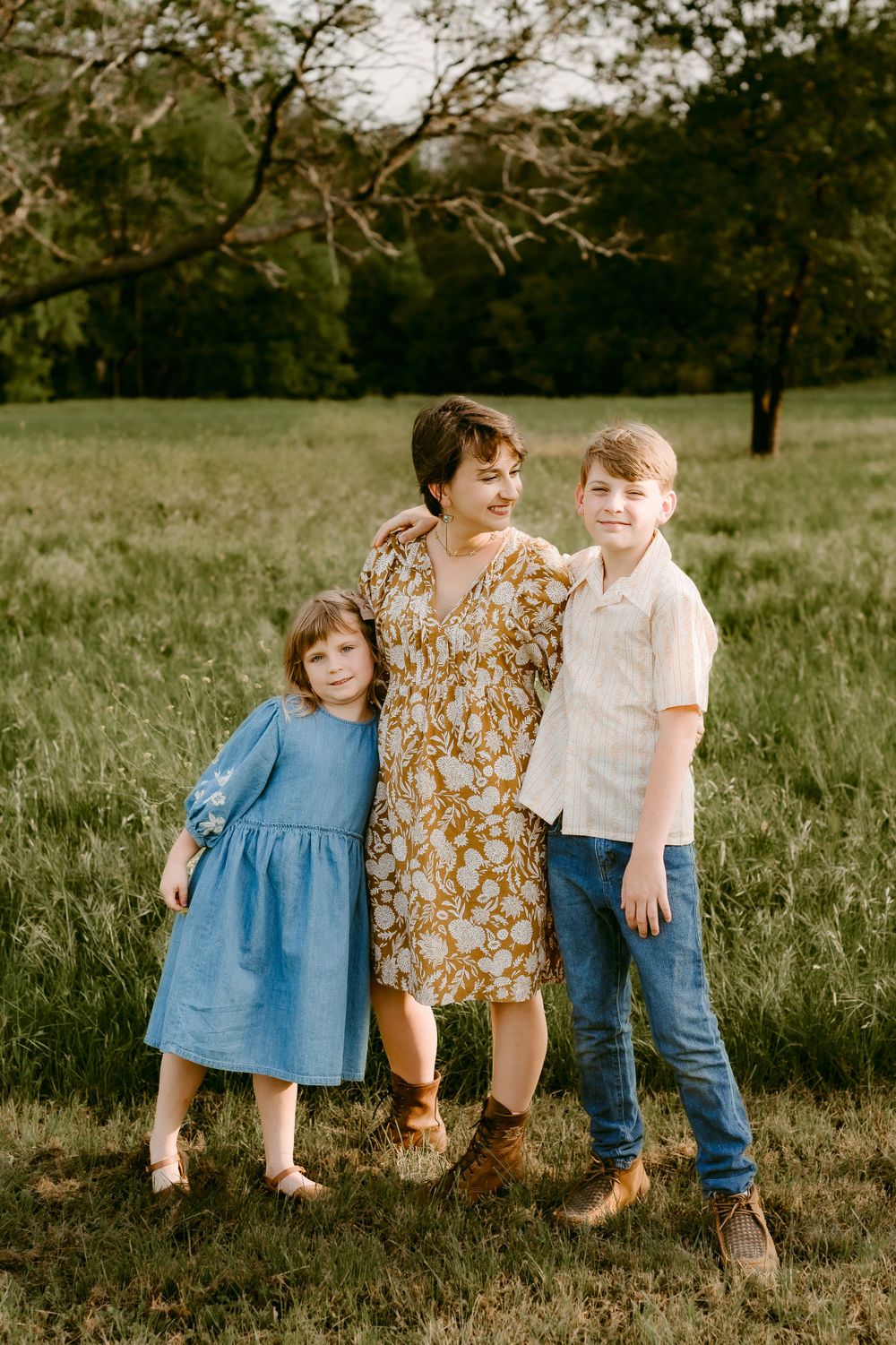 A family portrait in a grassy field with trees, featuring a floral dress and casual outfits in warm afternoon light.