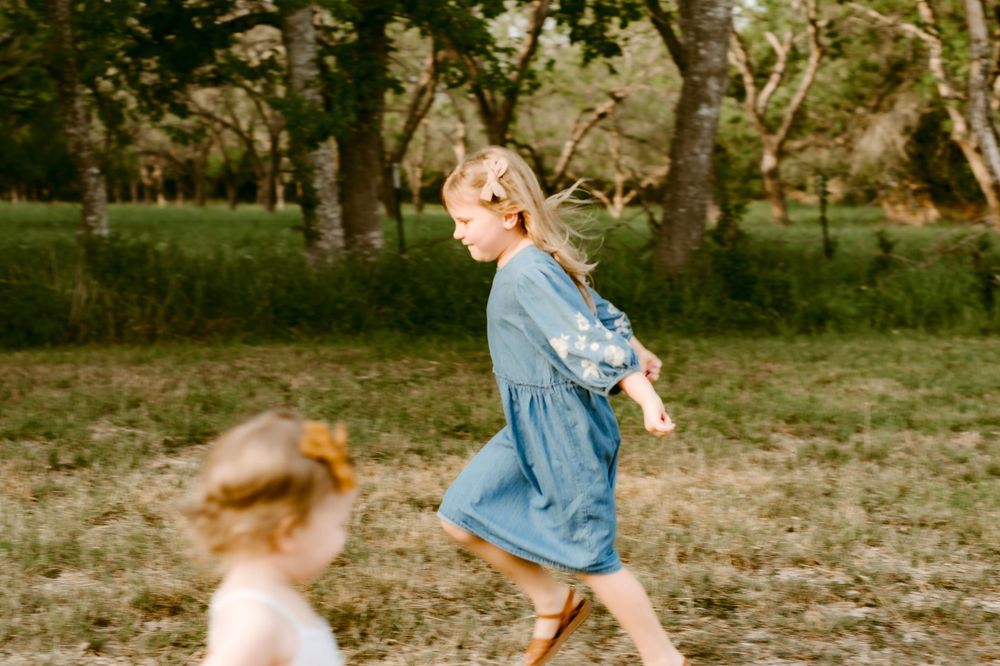 Two children running through a sunlit field wearing casual clothing.