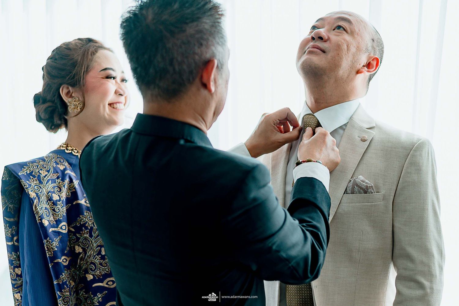 A helper adjusts the groom's tie while the bride watches with a smile.