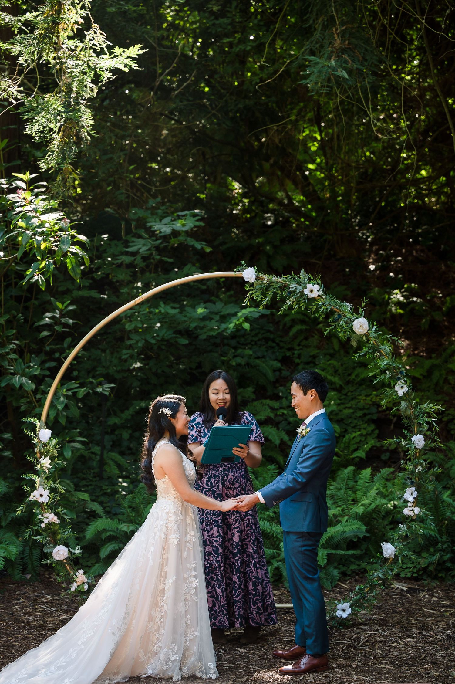 A wedding ceremony takes place under a floral arch in a secluded forest setting.
