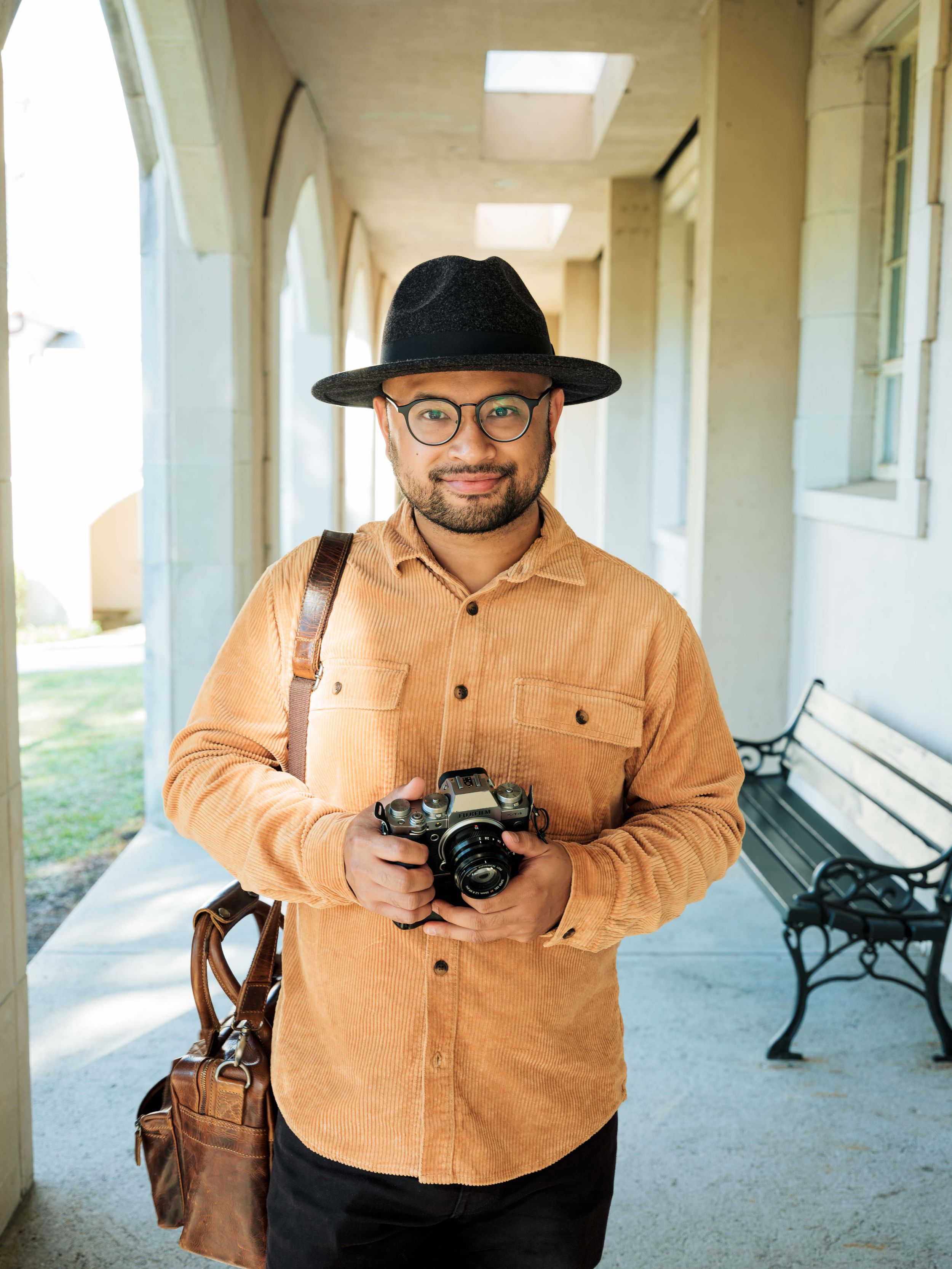 Photographer in tan shirt and black hat poses with camera in covered walkway.