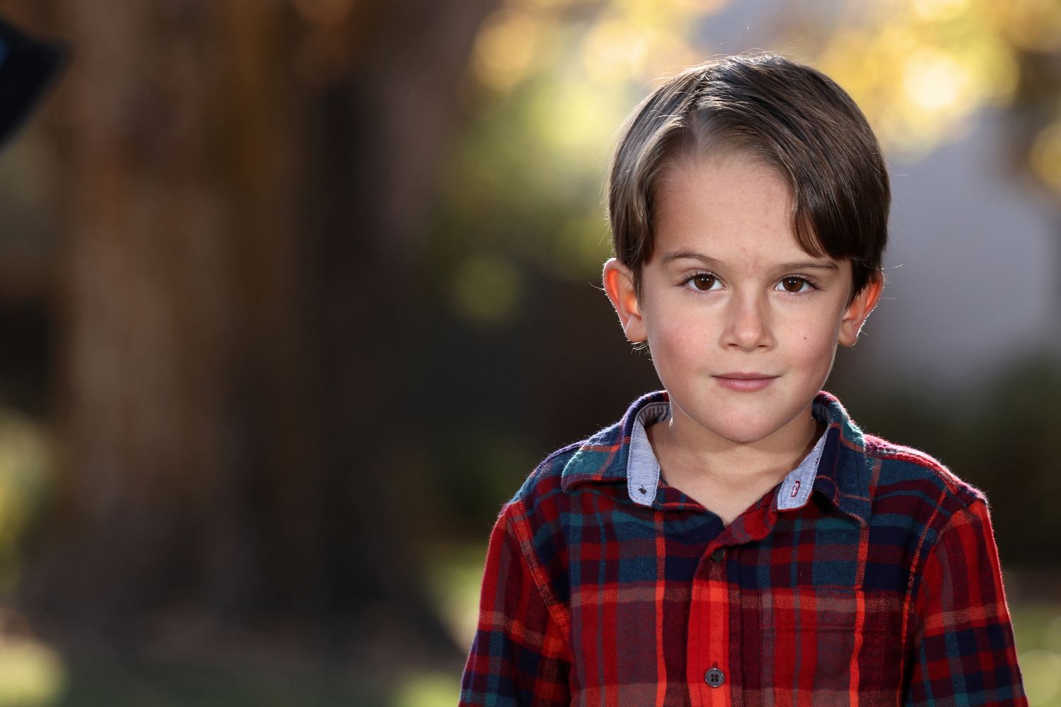 A young person in a red plaid shirt poses outdoors with soft bokeh background lighting.