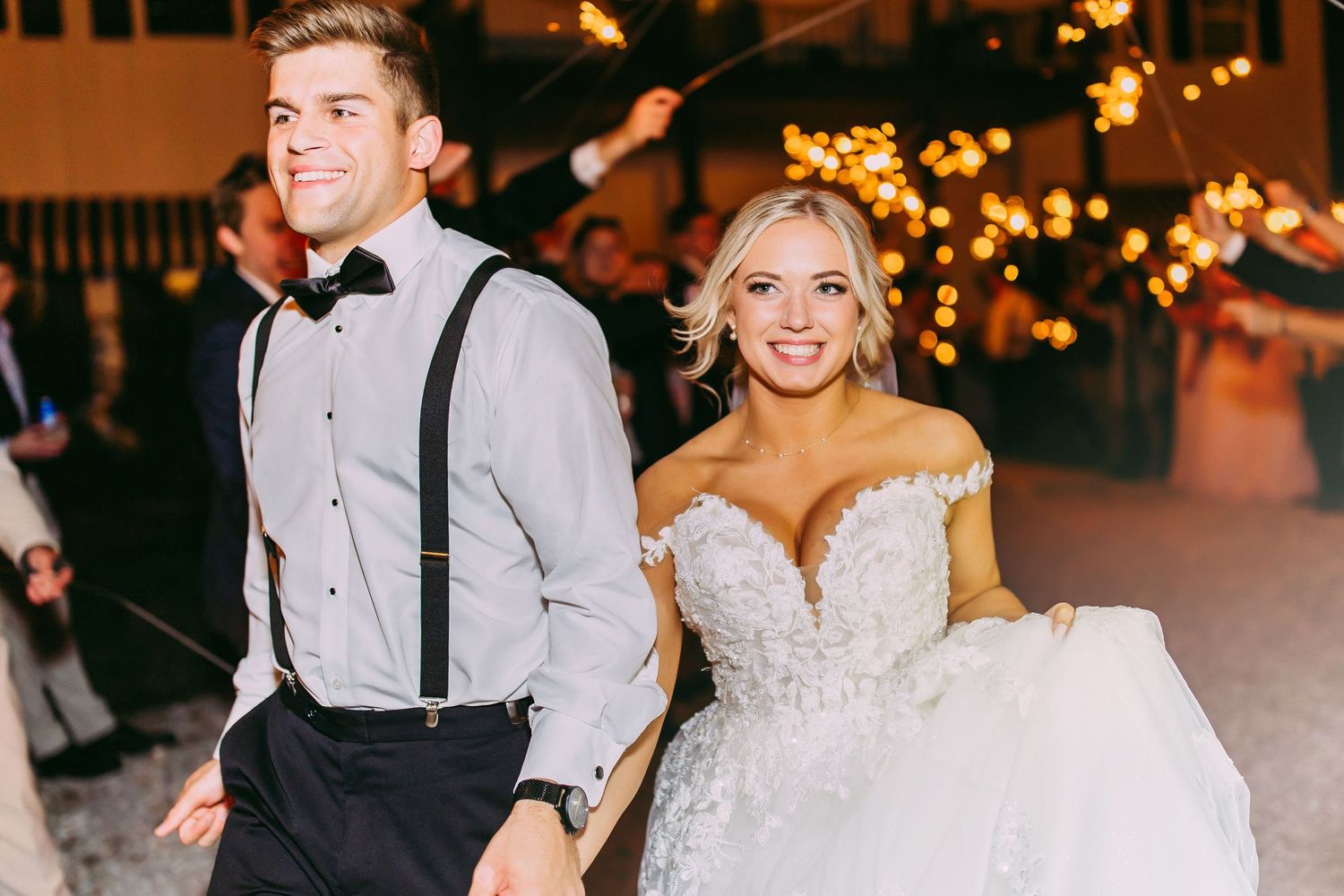 Elegantly dressed couple walks through reception venue with twinkling lights.