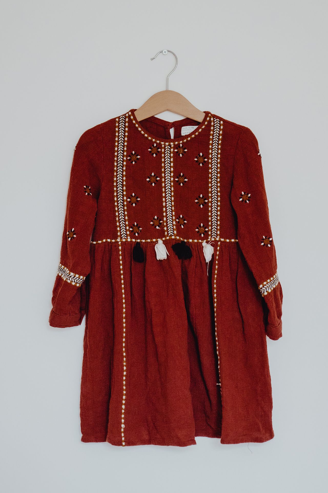 A red embroidered children's dress with tassel details hangs on a wooden hanger.