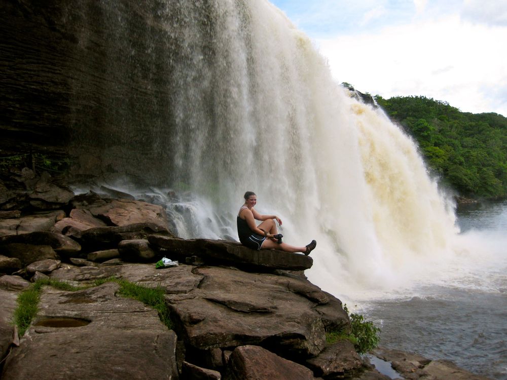 A dramatic waterfall flows over rocky cliffs while someone sits on the edge overlooking the scene.