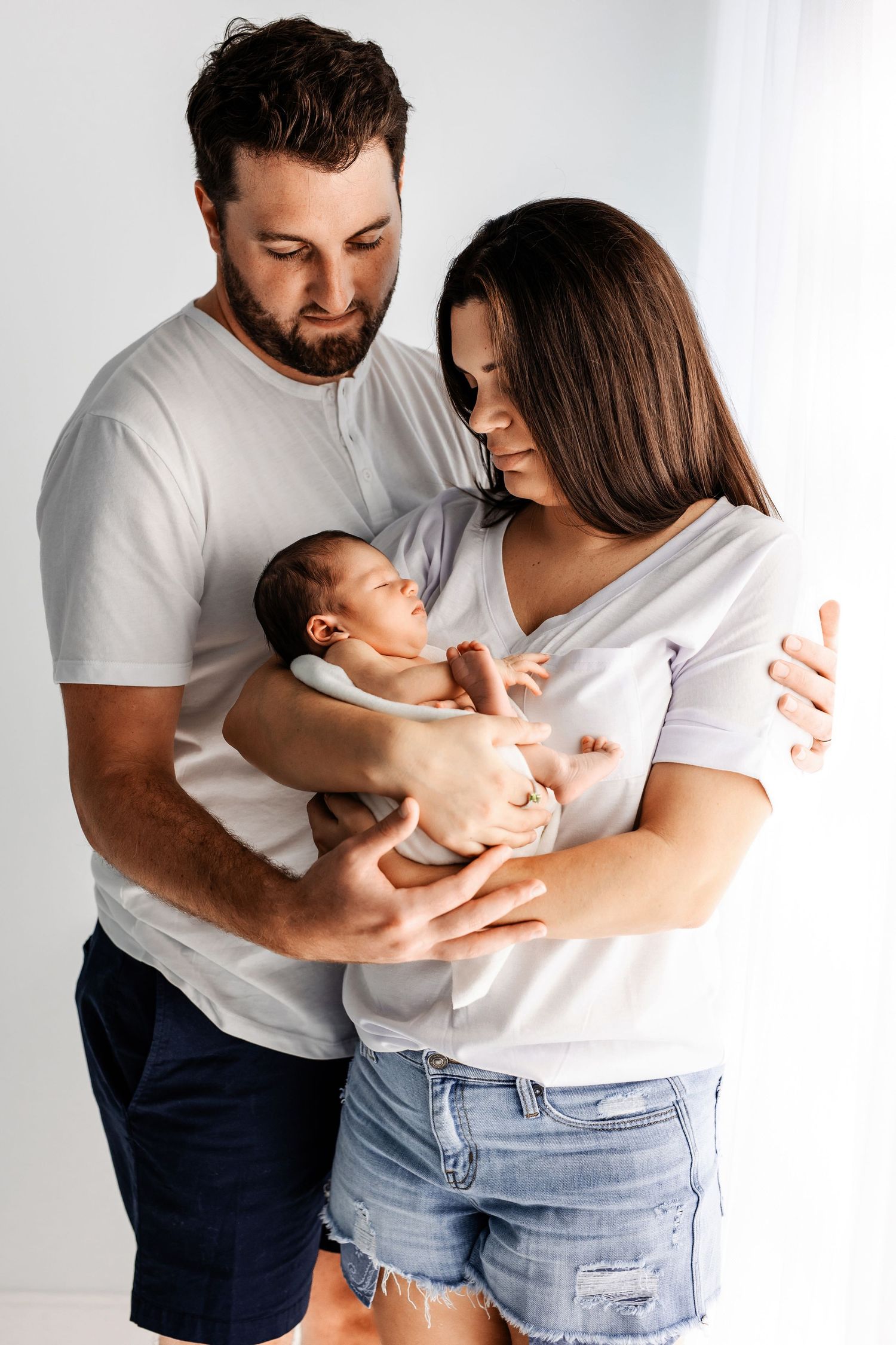 Couple in white shirts embraces while holding their newborn baby.