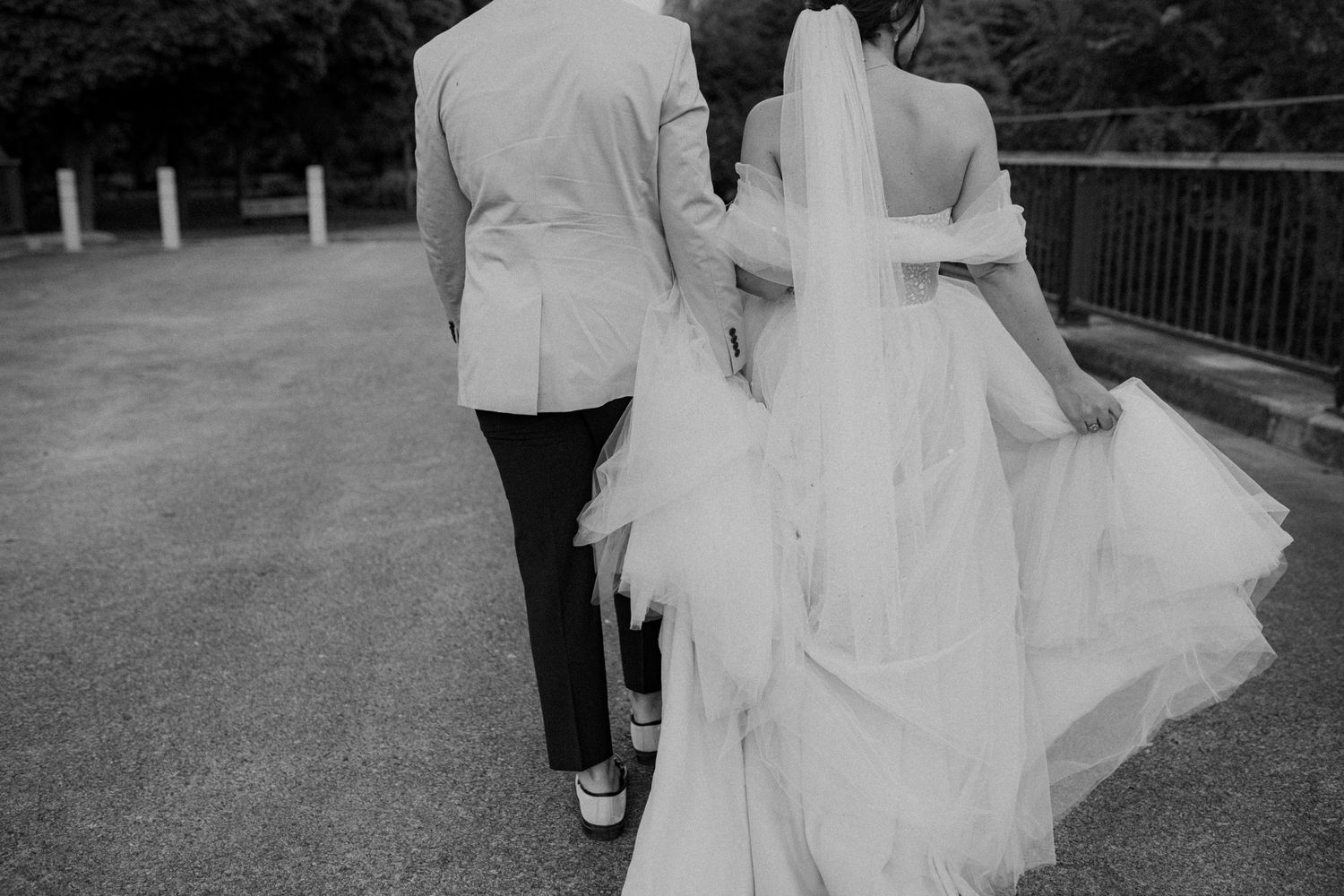 Black and white photo of bride and groom walking hand in hand, wedding dress with flowing veil trailing behind.