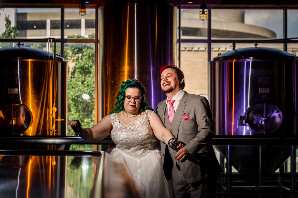 Couple poses in an industrial-style venue with copper brewing tanks in background.