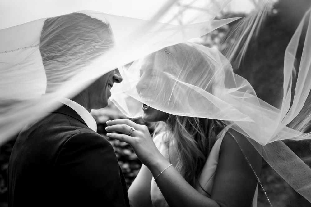 A couple embraces under flowing white veils against a bright background.